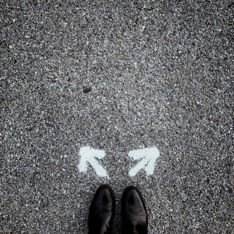Welchen Weg gehe ich? Schwarze Schuhe stehen auf einem Gehweg mit zwei weißen Pfeilen nach links und rechts.
